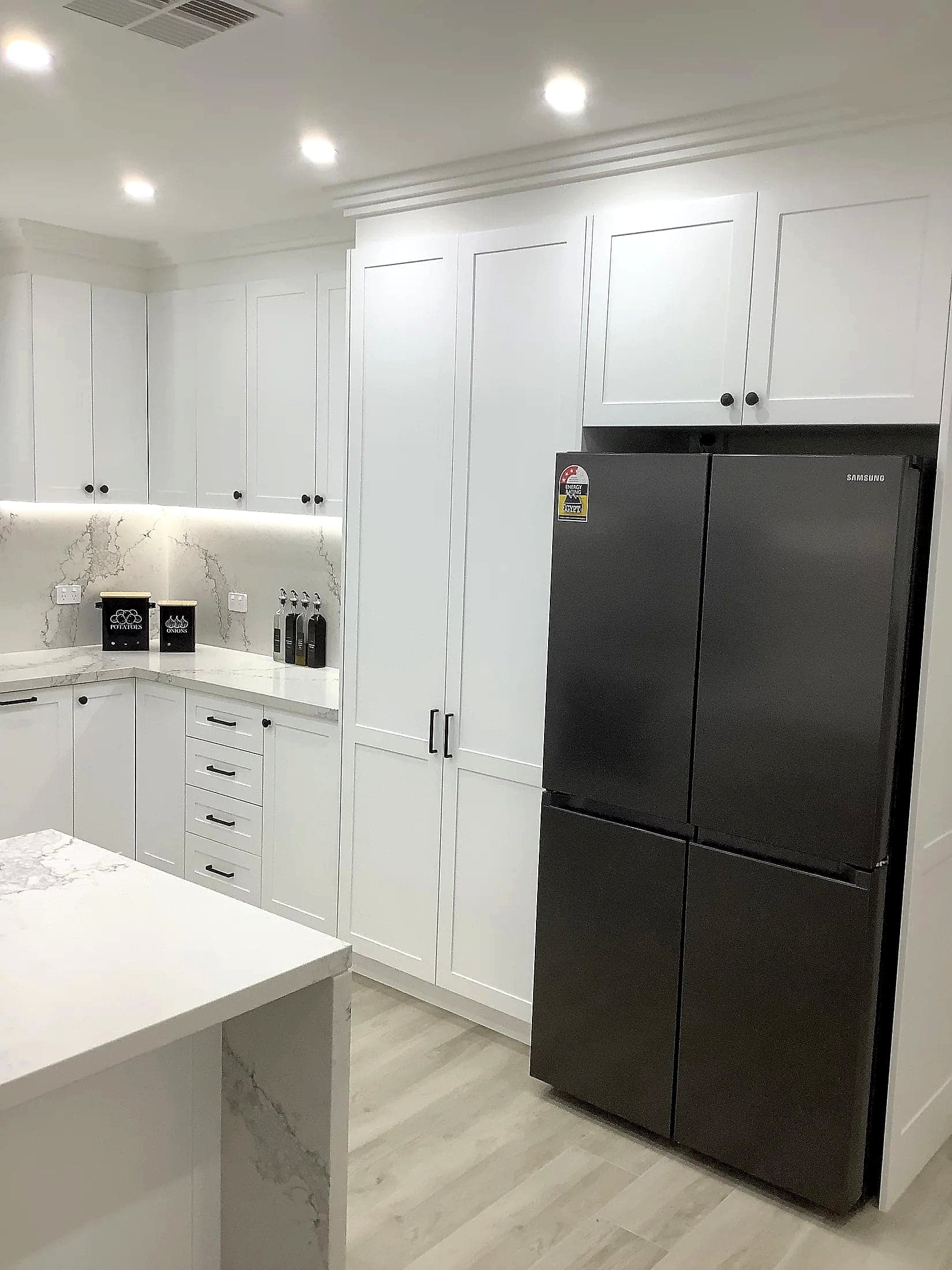 White shaker kitchen with marble-look benchtops, a tall pantry wall and a black French-door fridge by H&R Kitchens