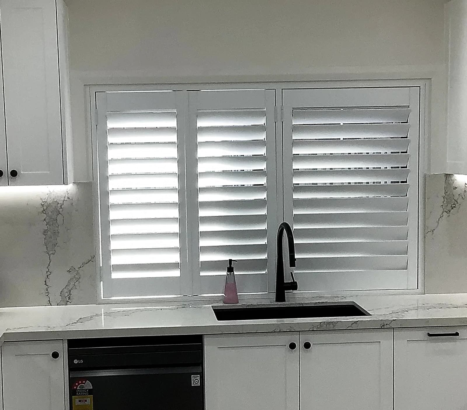 White shaker kitchen with a marble-look splashback and black sink beneath plantation shutters in Guildford