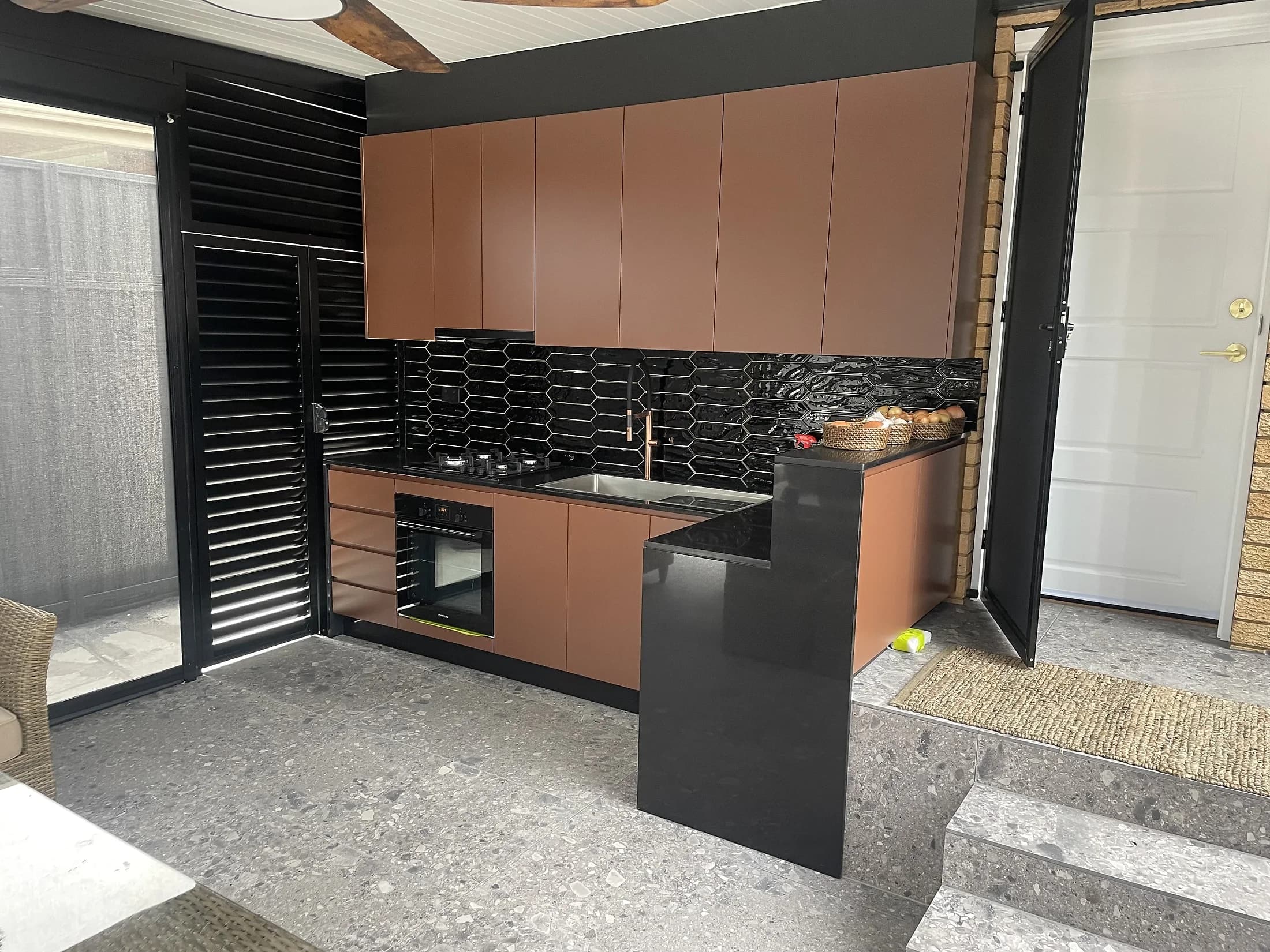 Covered alfresco kitchen with copper-toned cabinetry, dark benchtop and black tiled splashback by H&R Kitchens