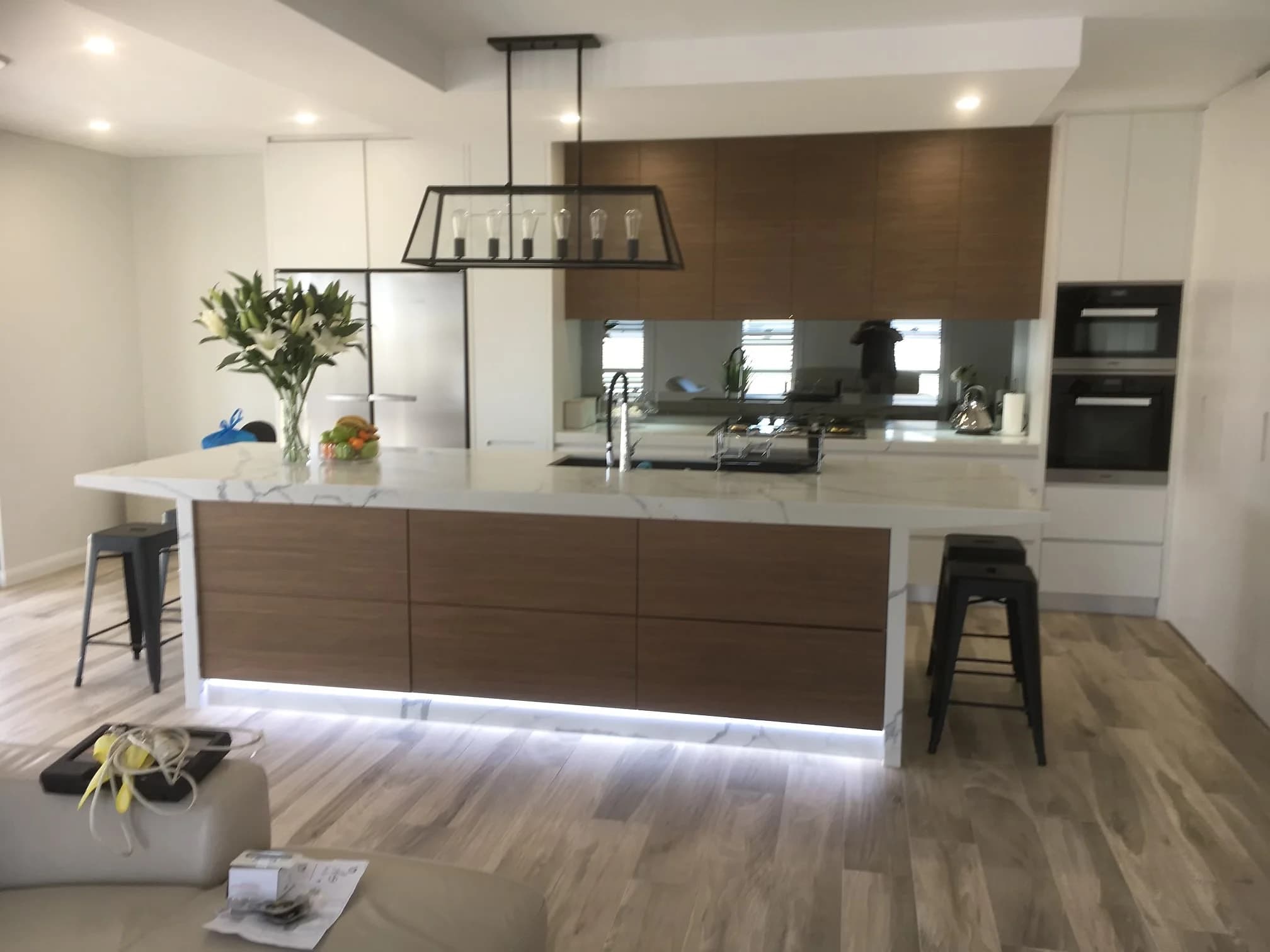 Open-plan kitchen with timber-look and white cabinetry, mirrored splashback and marble-look waterfall island by H&R Kitchens