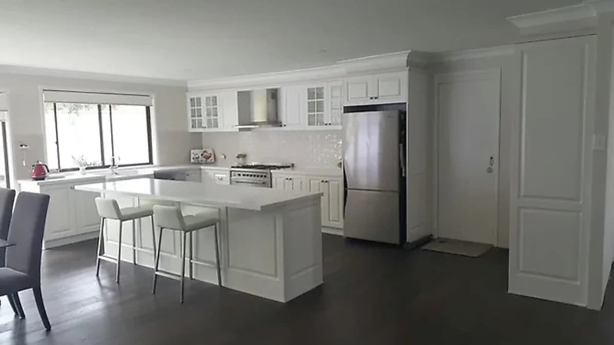 Traditional white kitchen with glass-front cabinetry, farmhouse sink and pale stone-look island by H&R Kitchens