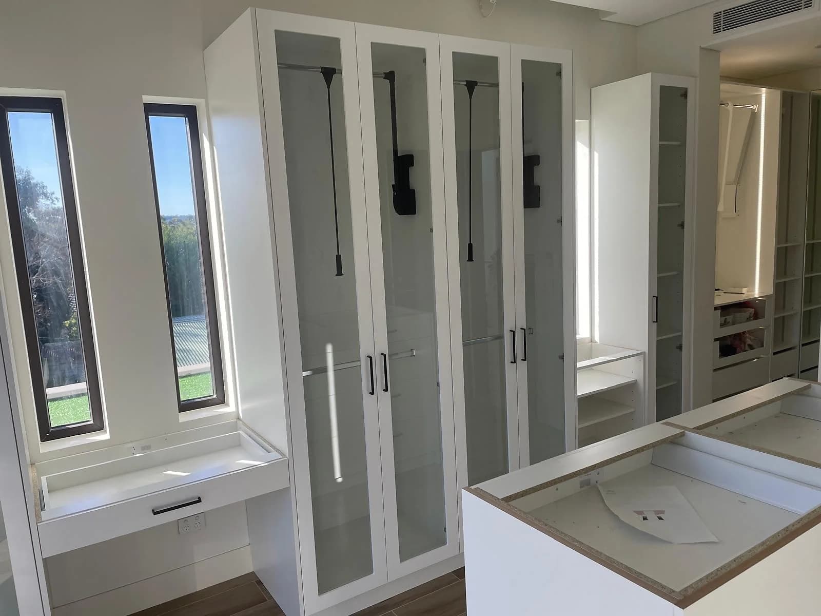 White walk-through wardrobe with glass-front cabinetry, hanging storage and a central drawer island by H&R Kitchens