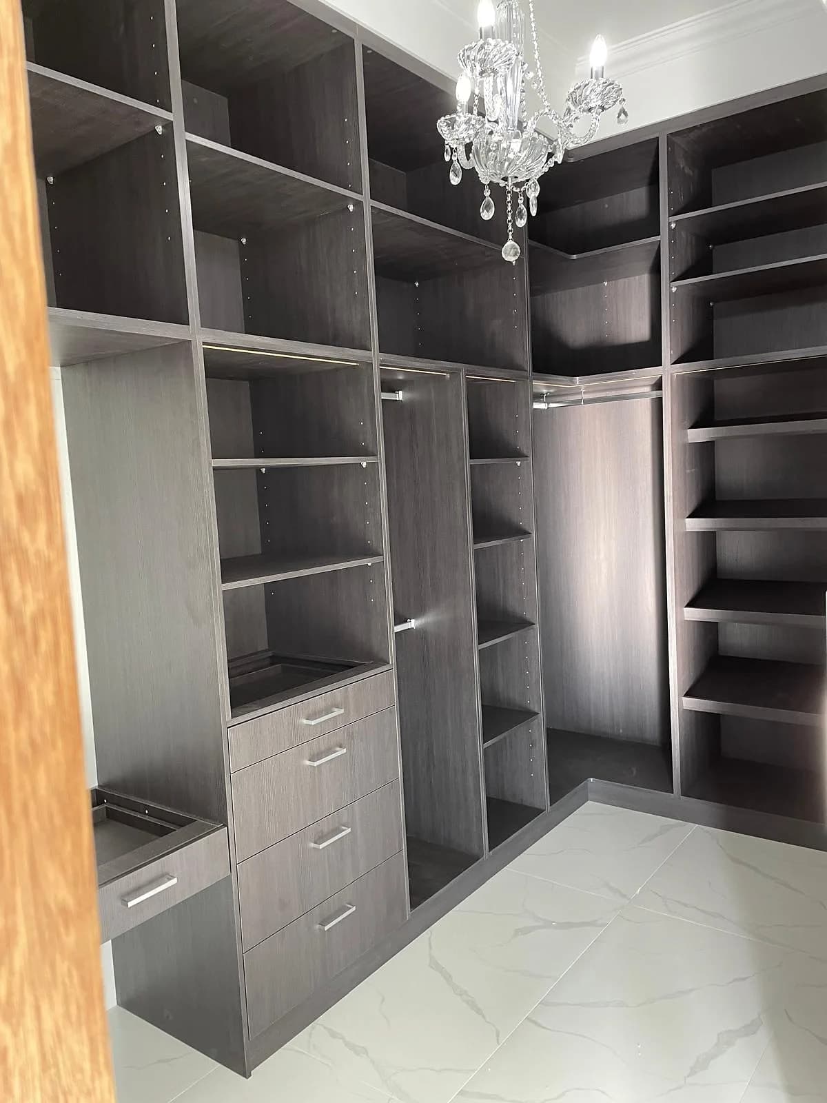Dark woodgrain walk-in wardrobe with open hanging, shoe storage and a crystal chandelier by H&R Kitchens