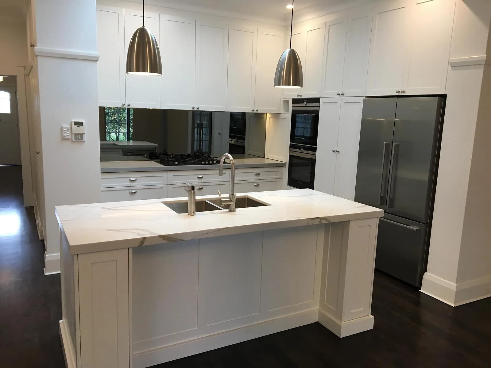 White shaker kitchen renovation with a marble-look waterfall island, mirror splashback and brushed-nickel pendants, by H&R Kitchens